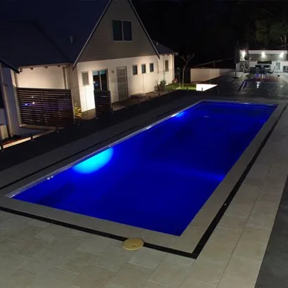 Platinum 9m fibreglass pool lit at night by underwater lights, with pale paving, frameless glass fencing and a white house beyond