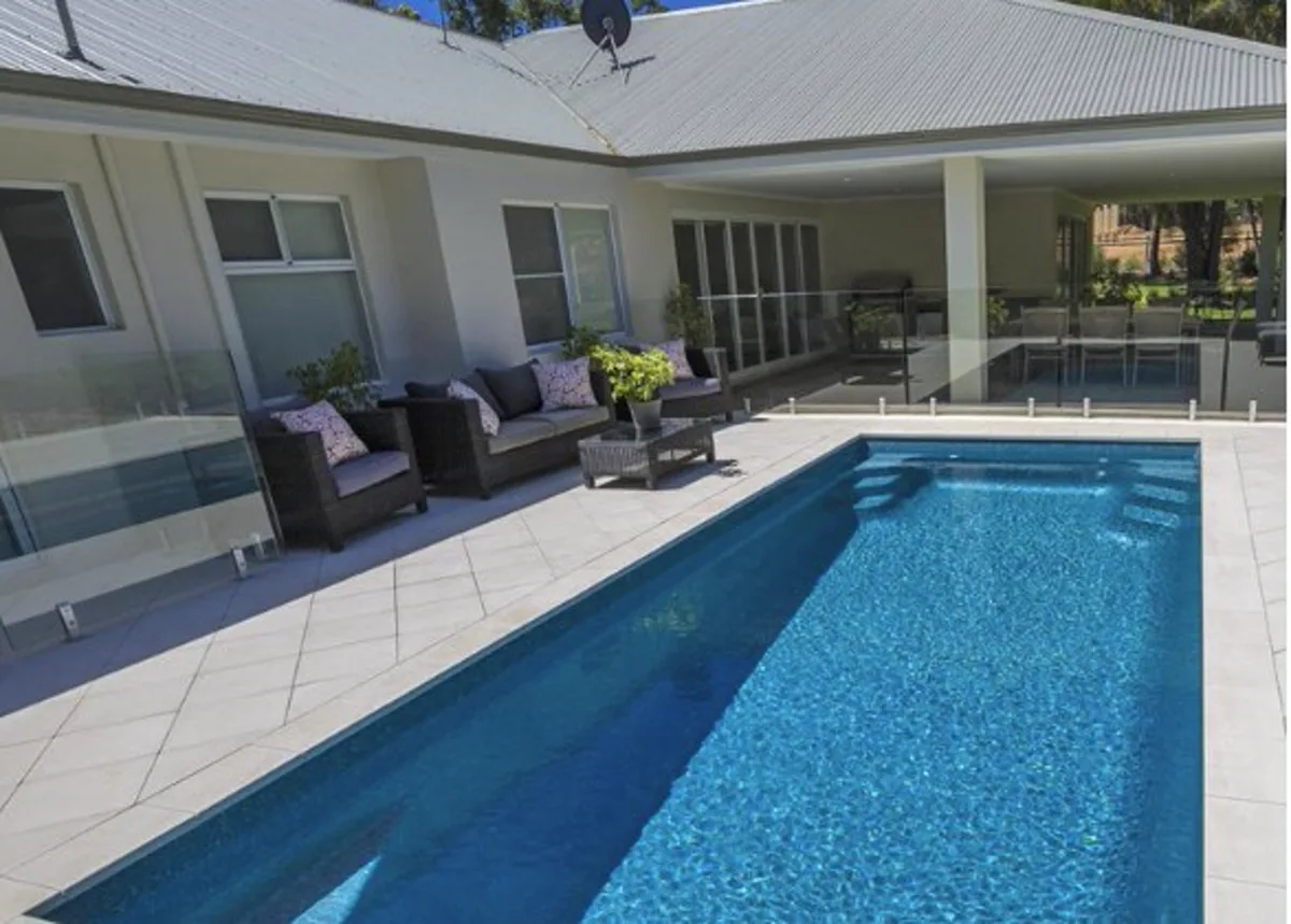 Platinum 11m fibreglass pool with tiered entry steps, pale paving and frameless glass fencing beside a modern home
